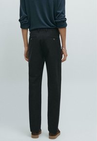 Schwarze Baumwollhosen mit geradem Schnitt, die zwei aufgesetzte Taschen mit Knöpfen und eine glatte Textur aufweisen. Kombiniert mit dunkelbraunen Schuhen.