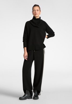 Mujer de pie con la mano en la cadera, viste un suéter negro holgado con cuello alto, pantalones anchos negros con rayas blancas en los lados y botas negras.