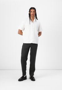 White short-sleeve button-up shirt made of lightweight fabric, paired with dark trousers and black shoes, standing against a plain background.