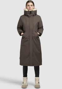 Brun lang foret jakke med en pelsforet hætte, to sidelommer og en lige skæring. Modellen har sorte leggings og kridhvide støvler på.