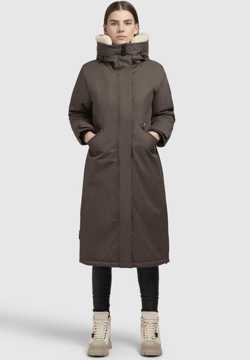 Brun lang foret jakke med en pelsforet hætte, to sidelommer og en lige skæring. Modellen har sorte leggings og kridhvide støvler på.