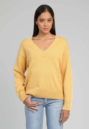 Femme aux longs cheveux bruns portant un pull jaune à col en V avec des détails blancs cousus sur les manches et un jean bleu clair, debout devant un fond gris.