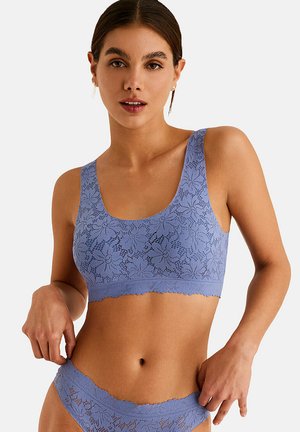 Femme portant un bralette en dentelle florale violet clair assorti et une culotte taille haute, posant avec les mains près des hanches sur un fond uni.