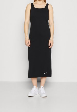 Kobieta ubrana w czarną, bezrękawnikową sukienkę midi z białym logo Nike w pobliżu dołu, w połączeniu z białymi trampkami, stojąca na jednolitym tle.