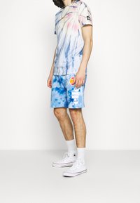 T-shirt tie-dye bleu et rose associée à un short bleu assorti orné d'un logo multicolore et de lettres blanches. Des baskets montantes blanches complètent la tenue.