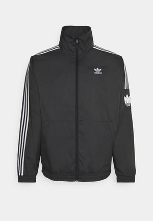 Osoba ubrana w czarny dres Adidas z białymi paskami i logo, białą koszulkę oraz białe tenisówki, stojąca na jasnym, prostym tle.
