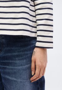 Χέρι με μικρό τατουάζ αράχνης κοντά στον καρπό, που φοράει μπλε τζιν και λευκό πουκάμισο με μαύρες οριζόντιες ρίγες.