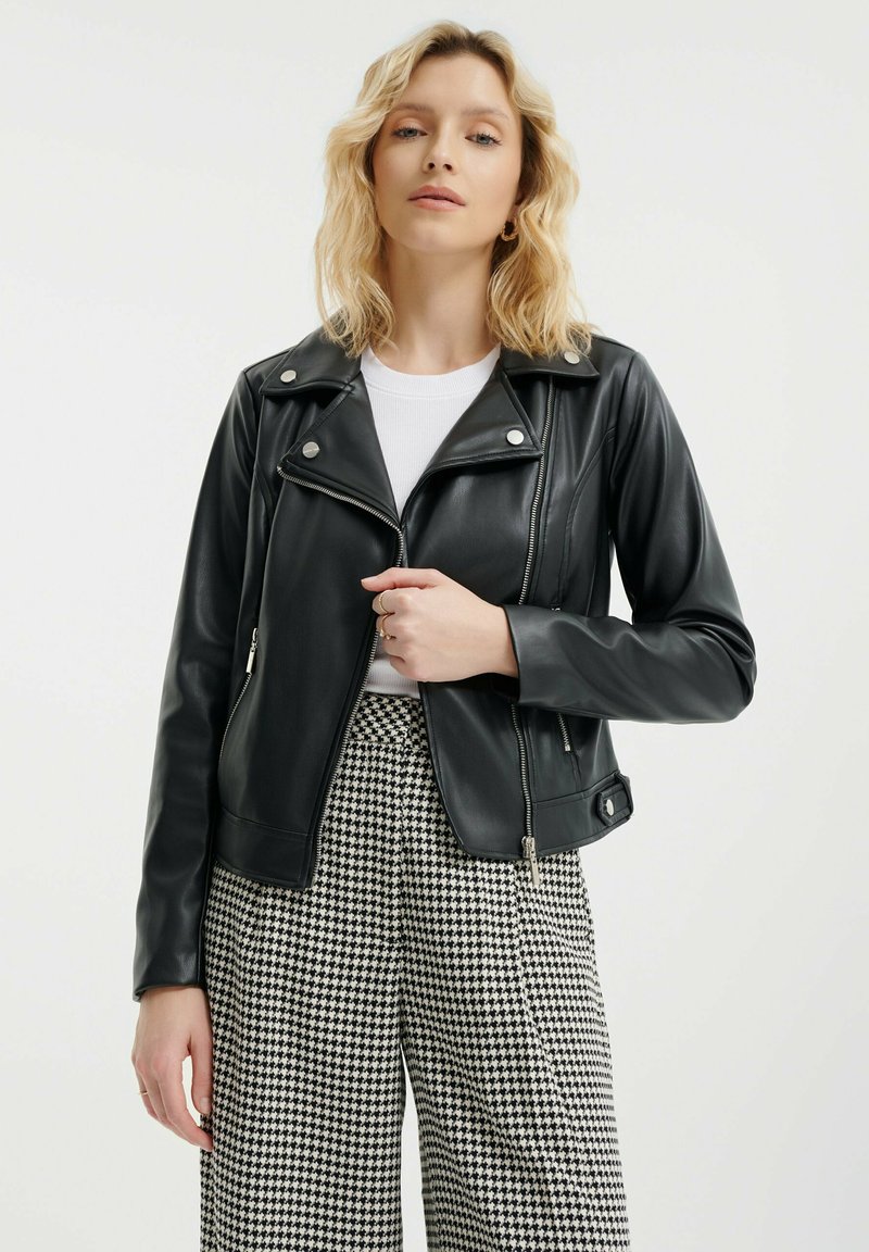Giacca da moto nera in ecopelle con hardware argentato, tasche laterali con zip e un design aderente, abbinata a pantaloni a scacchiera bianco e nero.