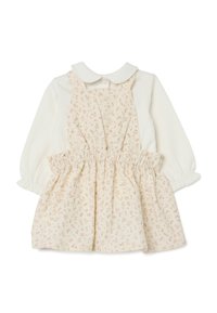 Witte lange mouwen top met een ronde kraag en een smocktail, gecombineerd met een beige jurk met bloemenpatroon en verstelbare schouderbanden.