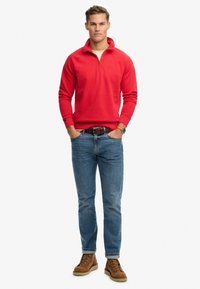 Suéter rojo con cuello con cremallera, combinado con jeans azules y botas de cordones marrones. El conjunto presenta un ajuste casual y relajado.