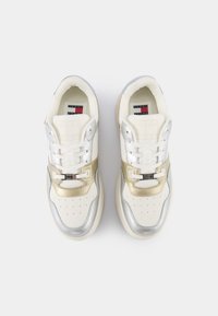 Paire de baskets blanches avec des accents métalliques argentés et dorés, lacets noués et logo Tommy Jeans visible sur les semelles intérieures.