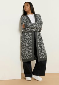 Lång grå cardigan med leopardmönster i svart och ljusgrått, stickad textur, öppen front, ribbade ärmslut, bärs över en vit T-shirt och svarta byxor.