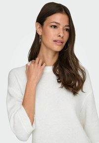 Pull en maille blanche avec un col rond et des manches trois-quarts, présentant une texture douce. Le modèle a des cheveux ondulés et des bijoux minimalistes.