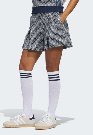 Persona con pantalones cortos azul marino con estampado, calcetas blancas hasta la rodilla con rayas negras y zapatillas Adidas beige con rayas blancas.