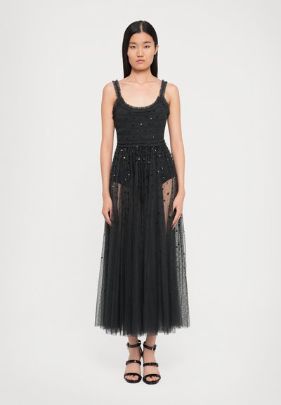 Woman stands wearing a sleeveless black tulle dress with sequins and ankle-strap black heels against a white background.