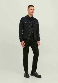Homem com uma jaqueta de ganga preta sobre uma camisa xadrez escura, calças pretas e botas pretas, posando contra um fundo claro e simples.
