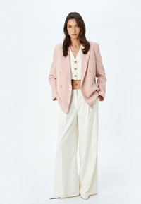 Blazer in Hellpink mit doppelter Knopfleiste, kombiniert mit einem weißen Crop-Top und weiten cremefarbenen Hosen, die goldene Knopfakzente aufweisen.