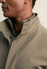 Beige Softshell-Jacke mit hohem Kragen, Frontreißverschluss und Druckknopfverschluss. Merkmale sind die glatte Textur und dezente Nähdetails.