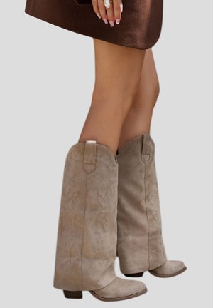 Jambes portant des bottes western hautes jusqu'aux genoux en daim beige, associées à une jupe marron, mains reposant près de l'ourlet de la jupe.