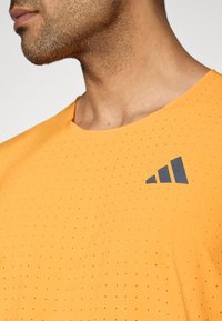 Πορτοκαλί κοντομάνικο πουκάμισο με διάτρητο σχέδιο και γκρι λογότυπο στο πάνω αριστερό μέρος του στήθους. Μαλακή υφή, στρογγυλή λαιμόκοψη.