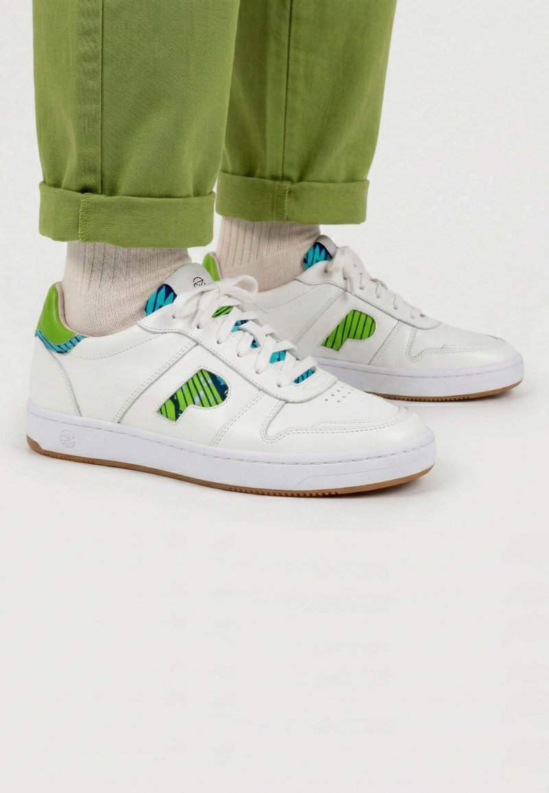 Baskets en cuir blanc avec des accents verts et bleus à motifs, portées avec des chaussettes beige et un pantalon vert retroussé sur un fond blanc.
