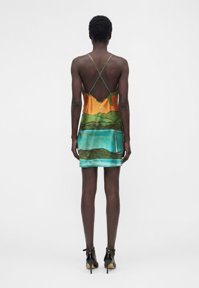 Γυναίκα που φοράει ένα μίνι φόρεμα χωρίς πλάτη με δυο σταυρωτές τιράντες, με πολύχρωμο σχέδιο τοπίου, στέκεται ξυπόλητη σε λευκό φόντο.