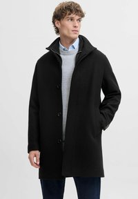 Must must coat kõrge kraega, esipaneeli nuppudega, küljetaskute ja siledat tekstuuri, seljas heledat halli jumpers ja sinised teksad.