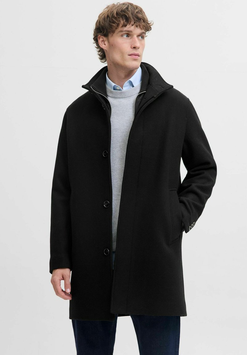 Must must coat kõrge kraega, esipaneeli nuppudega, küljetaskute ja siledat tekstuuri, seljas heledat halli jumpers ja sinised teksad.