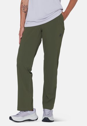 Osoba ubrana w oliwkowo-zielone, casualowe spodnie, jasnofioletowe trampki i jasnofioletową koszulę, stojąca z jedną ręką w kieszeni.