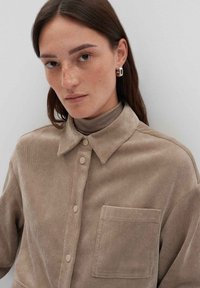 Camicia in velluto a coste marrone con colletto, chiusura frontale con bottoni e taschino sul petto. Texture liscia con sottili linee verticali.