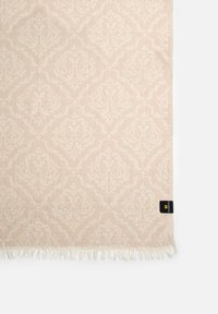 Vaalea beige kangas, jossa on hienovarainen damaskkuvio, frangeeratut reunat ja musta etiketti oikeassa alakulmassa.