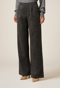 Weite Hosen aus strukturiertem schwarzem Stoff, mit hoher Taille und Frontfalten, kombiniert mit hautfarbenen High Heels.