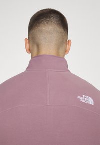 Fleecejakke i lyserød lilla med høj krave, der har hvidt logo detaljer på ryggen og flade sømme for et strømlinet design.