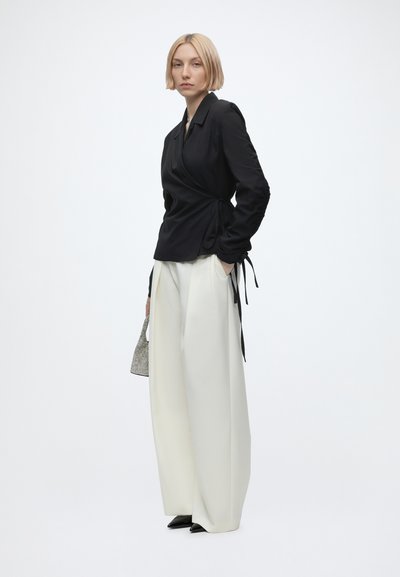 Femme aux cheveux blonds courts portant un chemisier noir cache-cœur et un pantalon large blanc, tenant un petit sac à main, debout sur un fond blanc.