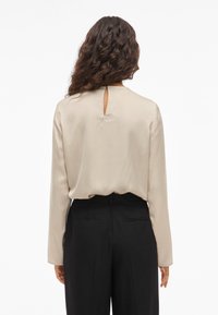 Μακρυμάνικη μπλούζα από σατέν υλικό σε μπεζ απόχρωση, με άν�οιγμα σε σχήμα κλειδαρότρυπας στην πλάτη και χαλαρή εφαρμογή, συνδυασμένη με μαύρο κομμένο παντελόνι.