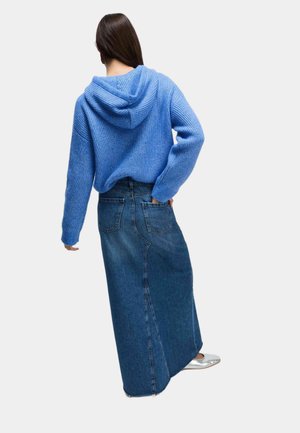 Sweat à capuche en maille bleue avec une finition texturée, associé à une longue jupe en denim à coupe droite avec des poches arrière. Chaussures plates argentées visibles.