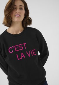 Musta huppari, jossa on näyttävä pinkki teksti "C'EST LA VIE", ribbaus kauluksessa ja pehmeä kangas.