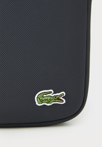 Schwarze, strukturierte Tasche mit einem grünen, gestickten Krokodil-Logo, das in Weiß umrandet ist. Kompaktes Design mit abgerundeten Kanten.