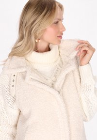 Cremefarbener Sherpa-Jacke mit texturiertem Finish, Reißverschlussfront und ribbeligem Rollkragen darunter. Mit Druckknopfdetails akzentuiert.