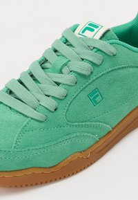 Zöld velúr sneaker gumitalppal, lapos fűzőkkel és logó címkével a nyelvén. Simma textúra és kerek orrtervezés.