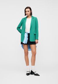 Femme portant un blazer vert, un haut blanc, une jupe noire, une chemise bleu clair nouée à la taille, des chaussettes blanches et des chaussures Mary Jane noires, debout avec confiance.