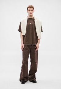 Brązowy t-shirt z okrągłym dekoltem z logo, w połączeniu z luźnymi brązowymi spodniami oraz kremowym swetrem z dzianiny zarzuconym na ramiona. Czarne buty.