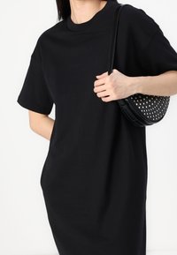 T-shirt noir oversize en coton doux. Il présente des manches courtes et un col rond. Un sac rond noir avec des clous métalliques est tenu.