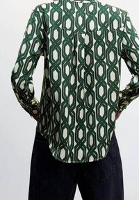 Chemise à manches longues avec des motifs géométriques verts en forme de chaîne, portée avec un pantalon foncé, vue de dos avec les mains sur les hanches.