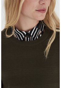 Blonde Frau, die einen olivgrünen strukturierten Pullover über einem schwarz-weiß-beigen, senkrecht gestreiften Hemd mit Kragen trägt.