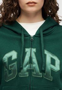 Ženska, oblečena v temno zeleno kapuco na zadrgo z velikimi svetlo zelenimi črkami "GAP" in belo majico spodaj, ki kaže njen spodnji del obraza in vrat.