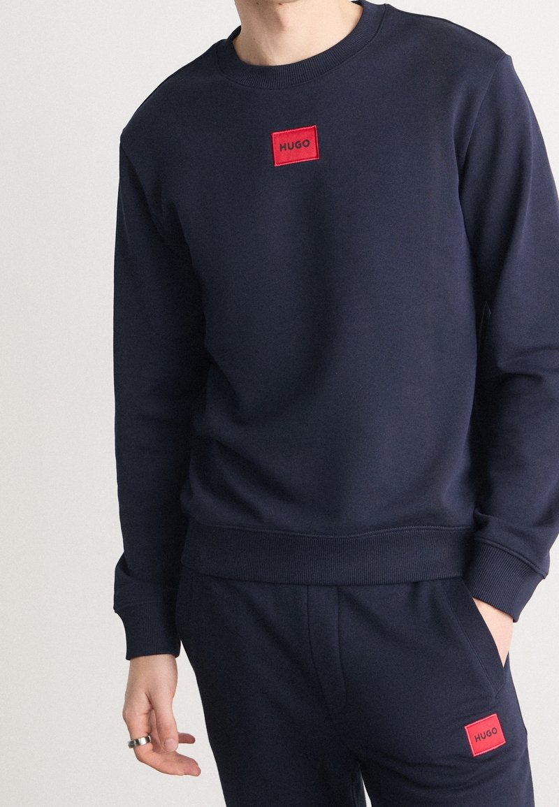 Ensemble sweat-shirt et pantalon bleu marine avec des patchs logo "HUGO" rouges sur la poitrine et la poche gauche, porté par une personne avec une main dans la poche.