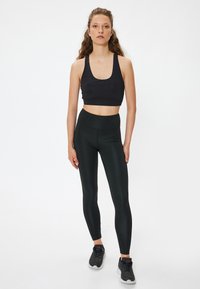 Soutien-gorge de sport noir avec bretelles larges, associé à un legging noir taille haute. Tissu lisse et extensible ; le modèle porte des baskets grises.