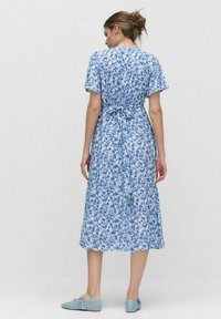 Vaaleansininen kukkamekko, jossa on lyhyet hihat, kiristetty vyötärö ja midi-pituus. Pehmeä kangas, jossa on virtaava muotoilu. Täydennetty sinisillä tasapohjaisilla kengillä.