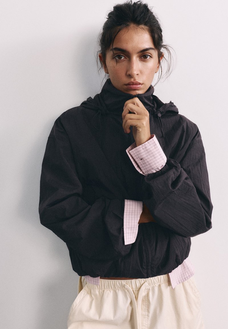 Jeune femme aux cheveux foncés portant une veste noire sur une chemise rose à carreaux et un pantalon beige, regardant directement avec une expression neutre.